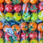 Verschiedene bunte Tomaten – rot, orange und gelb – sind in Plastikverpackungen mit kleinen Luftlöchern eingeschweißt. Die glänzende Folie reflektiert das Licht und zeigt die Tomaten in gleichmäßiger Anordnung, bereit für den Verkauf im Supermarkt.