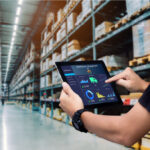 Person hält ein Tablet mit einem digitalen Dashboard für Lagerverwaltung in einem großen Warenlager mit hohen Regalen voller Kartons und Pakete.