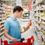 Ein Mann mit lockigen Haaren und blauem Polohemd steht mit einem Einkaufswagen in einem Supermarktgang. Er hält ein Glas mit grünen Oliven in der Hand und betrachtet das Etikett. Die Regale um ihn herum sind gefüllt mit Konservengläsern und Lebensmitteln.