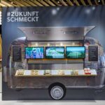 Messestand der BVE auf der Anuga 2021: Ein stilisierter Foodtruck mit der Aufschrift „#ZukunftSchmeckt“ zeigt auf drei Bildschirmen nachhaltige Projekte. Auf der Theke davor sind Lebensmittelproben und Produktinformationen ausgestellt. Der Stand ist modern gestaltet mit Holzelementen und aufmerksamkeitsstarkem Design.