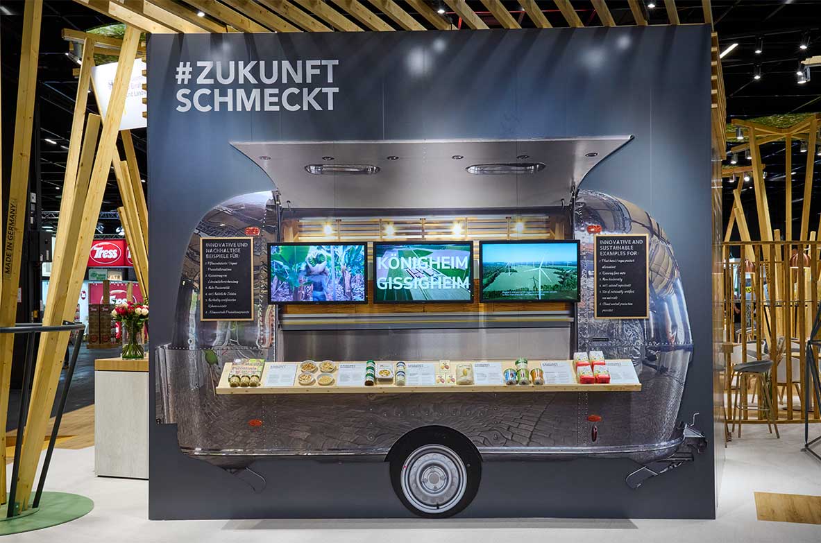 Messestand der BVE auf der Anuga 2021: Ein stilisierter Foodtruck mit der Aufschrift „#ZukunftSchmeckt“ zeigt auf drei Bildschirmen nachhaltige Projekte. Auf der Theke davor sind Lebensmittelproben und Produktinformationen ausgestellt. Der Stand ist modern gestaltet mit Holzelementen und aufmerksamkeitsstarkem Design.
