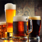 Vier verschiedene Biergläser mit unterschiedlichen Biersorten stehen auf einem rustikalen Holztisch. Ein hohes Weizenglas mit hellem, goldenem Bier und einer dichten Schaumkrone, ein klassischer Bierkrug mit bernsteinfarbenem Bier, ein dickwandiger Humpen mit rotbraunem Bier und ein Pint-Glas mit dunklem Bier und cremigem Schaum. Die warme Beleuchtung betont die leuchtenden Farben der Biere, während der Hintergrund aus dunklem Holz eine gemütliche Atmosphäre schafft.
