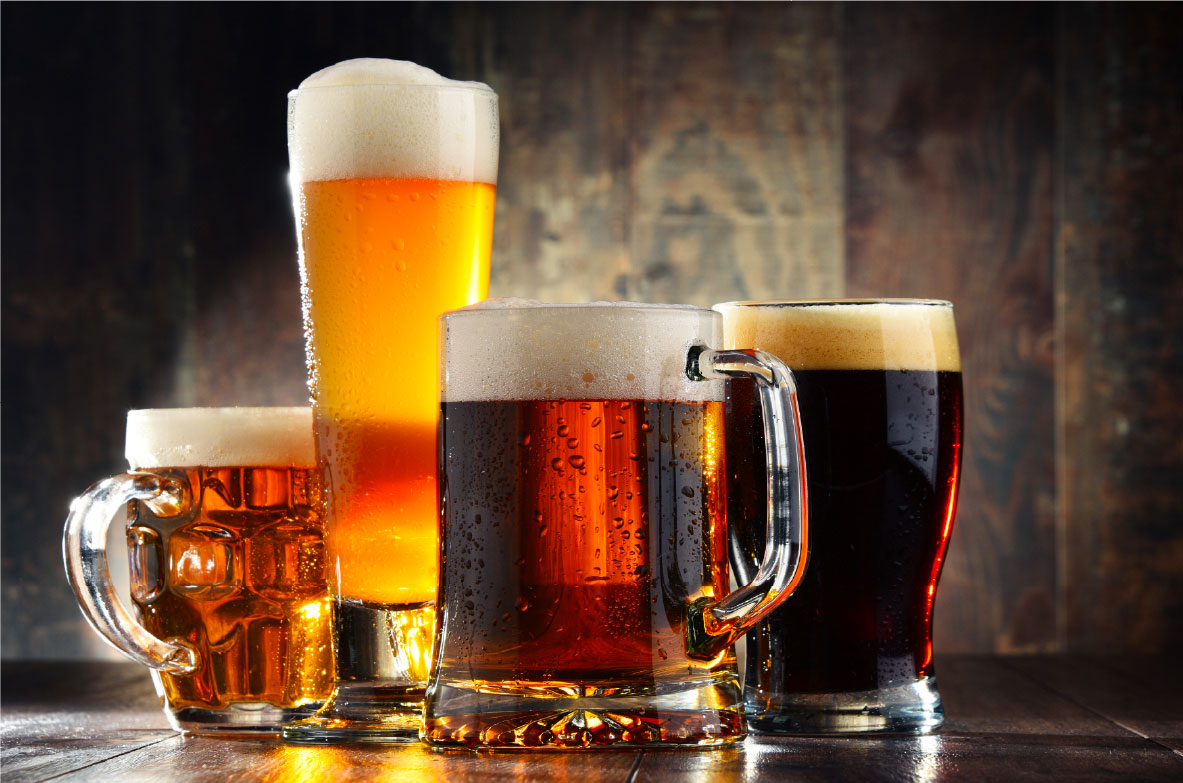 Vier verschiedene Biergläser mit unterschiedlichen Biersorten stehen auf einem rustikalen Holztisch. Ein hohes Weizenglas mit hellem, goldenem Bier und einer dichten Schaumkrone, ein klassischer Bierkrug mit bernsteinfarbenem Bier, ein dickwandiger Humpen mit rotbraunem Bier und ein Pint-Glas mit dunklem Bier und cremigem Schaum. Die warme Beleuchtung betont die leuchtenden Farben der Biere, während der Hintergrund aus dunklem Holz eine gemütliche Atmosphäre schafft.