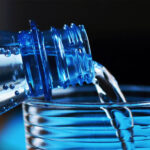 Nahaufnahme einer blauen Plastikflasche, aus der klares Wasser in ein geriffeltes Glas gegossen wird. Kleine Wasserperlen haften an der Flaschenöffnung. Der Hintergrund ist dunkel und unscharf.