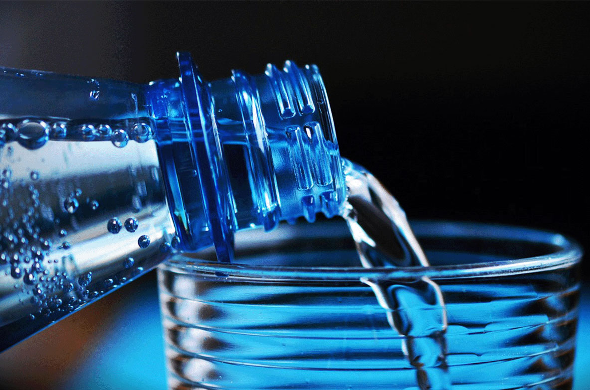 Nahaufnahme einer blauen Plastikflasche, aus der klares Wasser in ein geriffeltes Glas gegossen wird. Kleine Wasserperlen haften an der Flaschenöffnung. Der Hintergrund ist dunkel und unscharf.