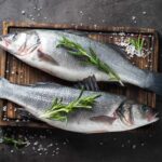 Zwei frische ganze Fische, dekoriert mit Rosmarinzweigen, liegen auf einem dunklen Holzbrett. Um sie herum sind grobes Meersalz und bunte Pfefferkörner auf einer dunklen Steinoberfläche verteilt.