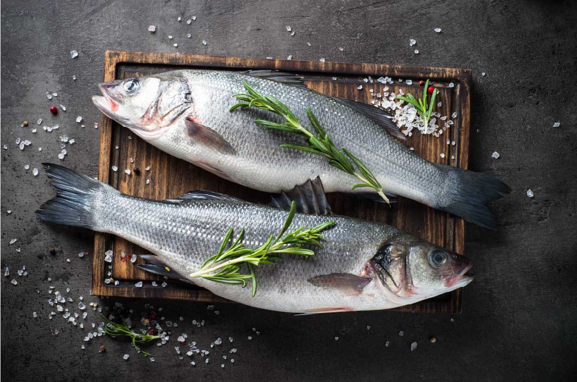 Zwei frische ganze Fische, dekoriert mit Rosmarinzweigen, liegen auf einem dunklen Holzbrett. Um sie herum sind grobes Meersalz und bunte Pfefferkörner auf einer dunklen Steinoberfläche verteilt.