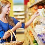 Eine junge Frau mit Einkaufskorb steht in einem Supermarkt vor einem Regal und greift lächelnd nach einer Packung Lebensmittel.