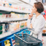 Junge Frau steht mit einem leeren Einkaufskorb im Kühlregal eines Supermarkts und betrachtet nachdenklich die Auswahl an Milch- und Molkereiprodukten. Sie trägt ein weißes Hemd und hat eine Hand nachdenklich ans Kinn geleg