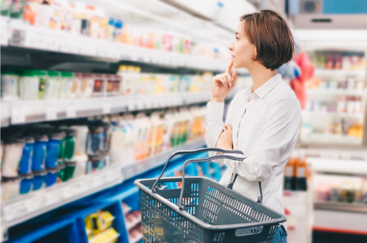 Junge Frau steht mit einem leeren Einkaufskorb im Kühlregal eines Supermarkts und betrachtet nachdenklich die Auswahl an Milch- und Molkereiprodukten. Sie trägt ein weißes Hemd und hat eine Hand nachdenklich ans Kinn geleg