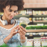 Junge Frau mit lockigen Haaren und Kopfhörern um den Hals steht in einem Supermarkt. Sie trägt eine gestreifte Bluse mit Latzhose und betrachtet aufmerksam die Verpackung eines grünen Smoothies. Im Hintergrund sind Regale mit Lebensmitteln und frischen Kräutern zu sehen.