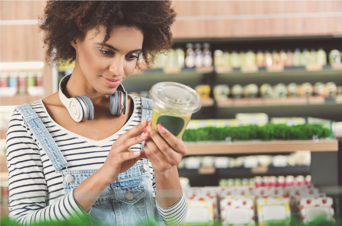 Junge Frau mit lockigen Haaren und Kopfhörern um den Hals steht in einem Supermarkt. Sie trägt eine gestreifte Bluse mit Latzhose und betrachtet aufmerksam die Verpackung eines grünen Smoothies. Im Hintergrund sind Regale mit Lebensmitteln und frischen Kräutern zu sehen.