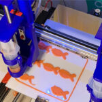 Ein 3D-Drucker stellt fischförmige Lebensmittel aus pflanzlichen Zutaten her. Die orangenen Fischattrappen werden schichtweise auf eine Druckplatte aufgetragen. Im Hintergrund ist das Logo von 'Revo Foods' zu sehen.