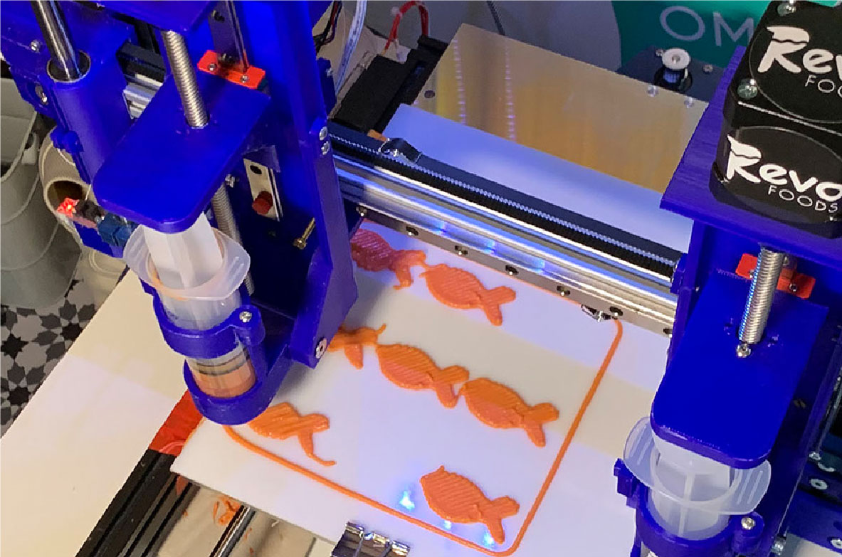 Ein 3D-Drucker stellt fischförmige Lebensmittel aus pflanzlichen Zutaten her. Die orangenen Fischattrappen werden schichtweise auf eine Druckplatte aufgetragen. Im Hintergrund ist das Logo von 'Revo Foods' zu sehen.