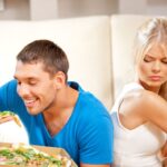 Ein Mann isst Pizza, seine Freundin einen Apfel. Sie schaut ablehnend auf ihren Mann.
