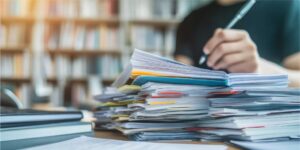 Ein hoher Stapel unsortierter Akten und Dokumente mit farbigen Markierungen und Notizzetteln liegt auf einem Schreibtisch. Im Hintergrund sitzt eine Person in schwarzer Kleidung und schreibt mit einem Stift, während eine große Bücherwand leicht verschwommen zu sehen ist.