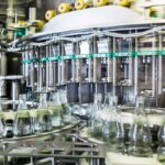 In einer hochmodernen Getränkeabfüllanlage fahren leere Glasflaschen auf einem Förderband unter präzise positionierte Düsen, aus denen eine klare Flüssigkeit abgefüllt wird. Die Anlage ist Teil eines industriellen Abfüllprozesses in der Lebensmittel- oder Getränkeindustrie und steht für Effizienz, Hygiene und moderne Produktionstechnologie.