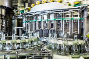 In einer hochmodernen Getränkeabfüllanlage fahren leere Glasflaschen auf einem Förderband unter präzise positionierte Düsen, aus denen eine klare Flüssigkeit abgefüllt wird. Die Anlage ist Teil eines industriellen Abfüllprozesses in der Lebensmittel- oder Getränkeindustrie und steht für Effizienz, Hygiene und moderne Produktionstechnologie.