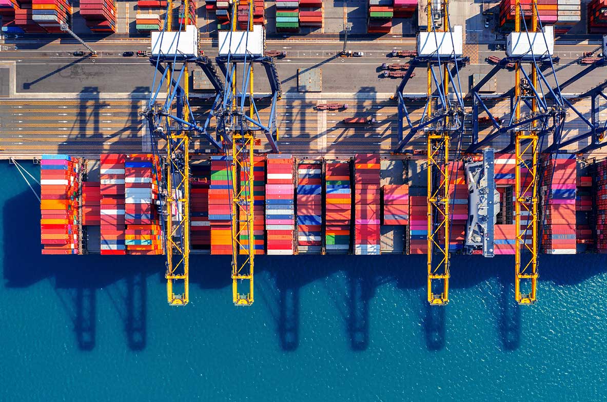 Luftaufnahme eines Containerhafens mit farbenfrohen Schiffscontainern, gelben Verladekränen und tiefblauem Wasser.