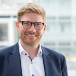 Tobias Koppitz im Porträt – lächelnd, mit Brille und Business-Outfit.