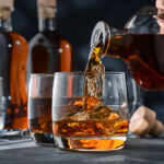 Whiskey wird aus einer Karaffe in ein Glas eingeschenkt, im Hintergrund stehen weitere Flaschen und Gläser.