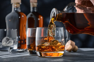 Whiskey wird aus einer Karaffe in ein Glas eingeschenkt, im Hintergrund stehen weitere Flaschen und Gläser.