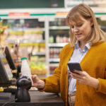 Frau scannt ein Produkt an der Supermarktkasse und hält dabei ein Smartphone in der Hand