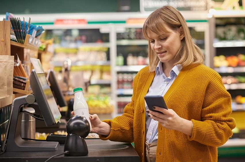 Frau scannt ein Produkt an der Supermarktkasse und hält dabei ein Smartphone in der Hand