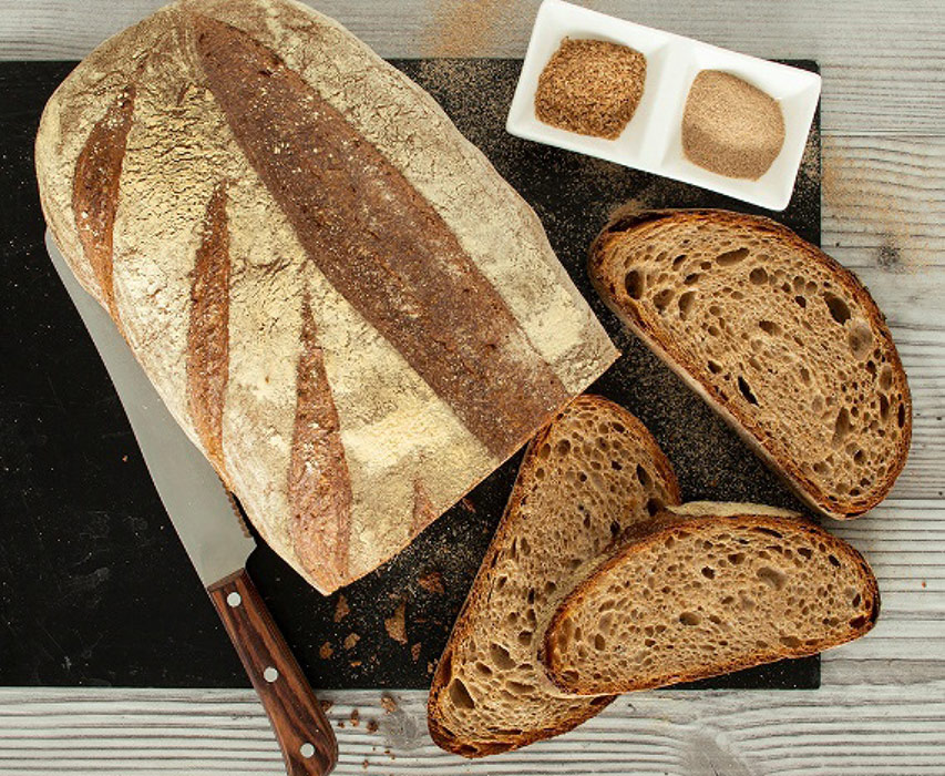 Aufgeschnittenes Laibbrot mit Kruste und zwei Schalen mit Getreidebestandteilen auf einem Tisch.
