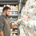 Ein Mann steht im Supermarkt vor einem Kühlregal und prüft aufmerksam das Etikett einer Milchflasche – Symbolbild für bewussten Einkauf und Preisvergleich.