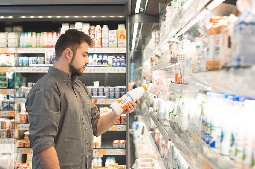 Ein Mann steht im Supermarkt vor einem Kühlregal und prüft aufmerksam das Etikett einer Milchflasche – Symbolbild für bewussten Einkauf und Preisvergleich.