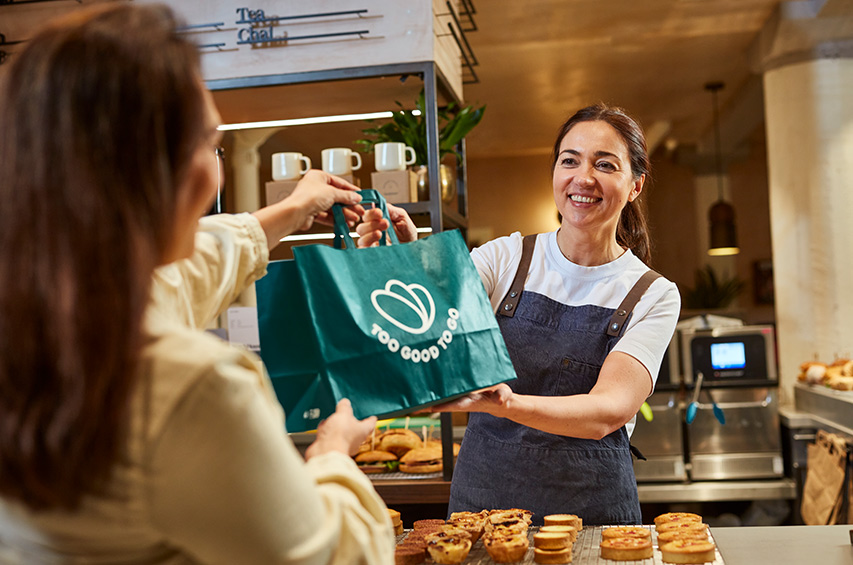 Frau übergibt einer Kundin eine Too Good To Go Tasche mit Lebensmitteln.