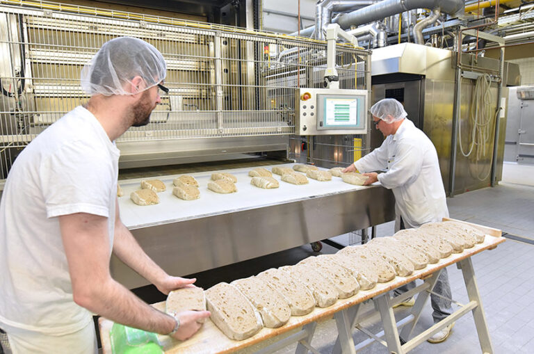 Mitarbeiter in einer Bäckerei bereiten Brotlaibe auf einem Förderband vor; im Hintergrund moderne Backtechnik.