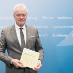 Bundesernährungsminister Alois Rainer hält den Ernährungsreport 2025 vor einer blauen Wand mit Ministeriumsbegriffen hoch.