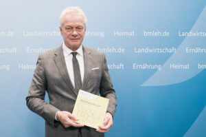 Bundesernährungsminister Alois Rainer hält den Ernährungsreport 2025 vor einer blauen Wand mit Ministeriumsbegriffen hoch.