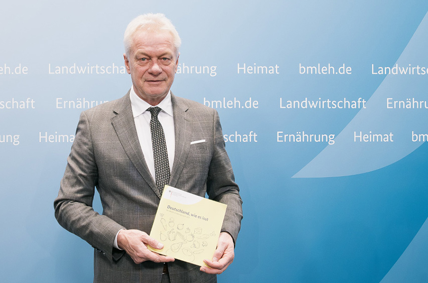 Bundesernährungsminister Alois Rainer hält den Ernährungsreport 2025 vor einer blauen Wand mit Ministeriumsbegriffen hoch.