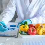 Person im weißen Laborkittel arbeitet im Labor mit frischem Obst und Gemüse und notiert Daten auf einem Klemmbrett.