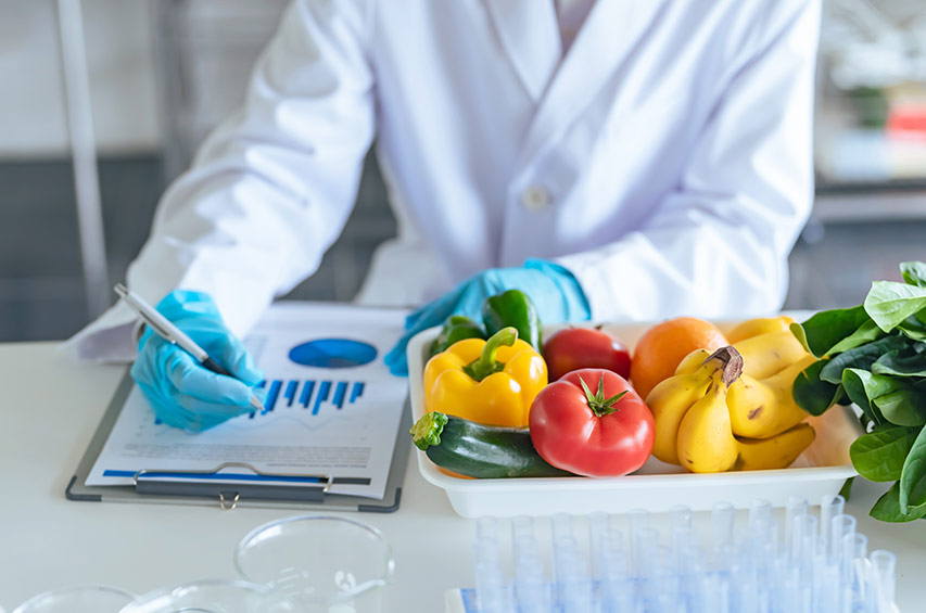 Person im weißen Laborkittel arbeitet im Labor mit frischem Obst und Gemüse und notiert Daten auf einem Klemmbrett.