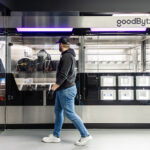 Ein Mitarbeitender steht vor der GoodBytz-Kochstation, in der Roboterarme Zutaten verarbeiten und Gerichte zubereiten.