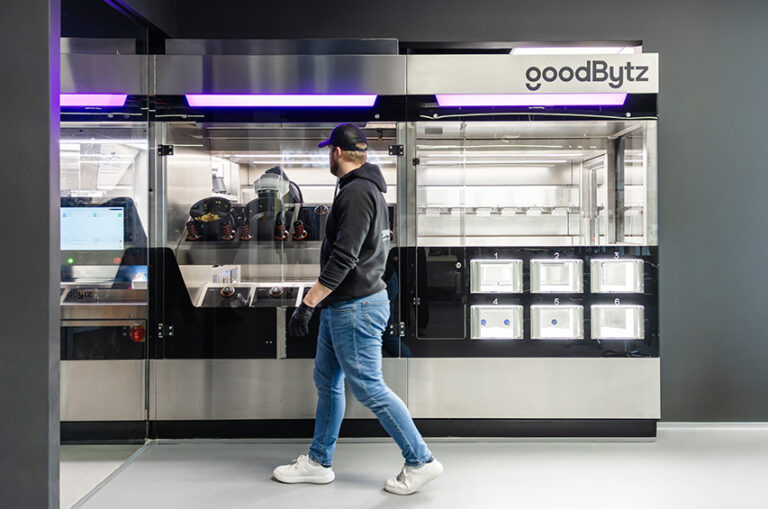 Ein Mitarbeitender steht vor der GoodBytz-Kochstation, in der Roboterarme Zutaten verarbeiten und Gerichte zubereiten.