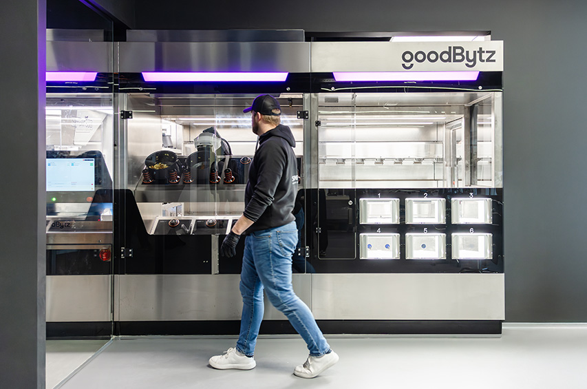 Ein Mitarbeitender steht vor der GoodBytz-Kochstation, in der Roboterarme Zutaten verarbeiten und Gerichte zubereiten.