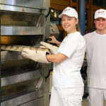Zwei Auszubildende in weißer Arbeitskleidung stehen in einer Backstube. Eine Person schiebt mit hitzebeständigen Handschuhen ein Blech in einen industriellen Etagenbackofen, eine zweite Person steht daneben.