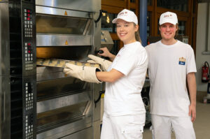 Zwei Auszubildende in weißer Arbeitskleidung stehen in einer Backstube. Eine Person schiebt mit hitzebeständigen Handschuhen ein Blech in einen industriellen Etagenbackofen, eine zweite Person steht daneben.