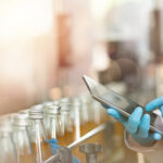 Mitarbeiter in einer Getränkeproduktion prüft Produktionsdaten auf einem Tablet zwischen abgefüllten Glasflaschen.