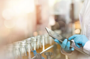 Mitarbeiter in einer Getränkeproduktion prüft Produktionsdaten auf einem Tablet zwischen abgefüllten Glasflaschen.