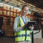 Mitarbeiterin in einer Lagerhalle prüft Liefer- und Bestandsdaten auf einem Tablet zwischen Hochregalen mit Waren.