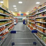 Blick durch einen Supermarktgang aus der Perspektive eines Einkaufswagens, beidseitig dicht gefüllte Regale mit Lebensmitteln