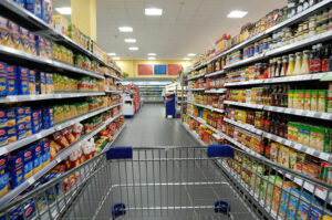 Blick durch einen Supermarktgang aus der Perspektive eines Einkaufswagens, beidseitig dicht gefüllte Regale mit Lebensmitteln