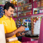 Ladenbesitzer in einem indischen Kirana-Shop verarbeitet eine Kartenzahlung, im Hintergrund vollbepackte Regale mit Lebensmitteln und Snacks