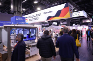 Besucherinnen und Besucher auf einem gut besuchten Messestand der Bundesvereinigung der Deutschen Ernährungsindustrie auf der Anuga. Über dem Stand hängt ein großes Banner mit der Aufschrift „Food & Drink Industries Germany“. Im Vordergrund ist moderne Lebensmitteltechnologie zu sehen, während zahlreiche Fachbesucher durch die Messehalle gehen. Das Bild steht für Export, Innovation und die internationale Präsenz der deutschen Ernährungsindustrie.